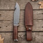 Damascus gut hook knife with stacked leather handle laid flat on oak planks next to its brown leather sheath