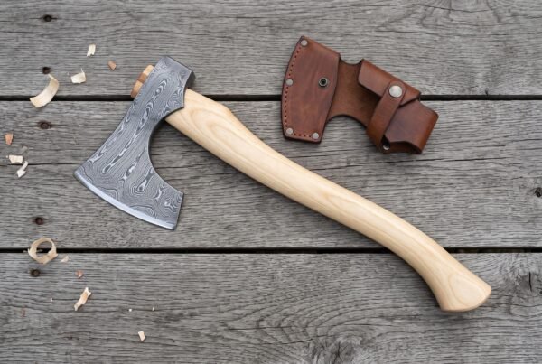Damascus bearded axe with long birch handle on grey planks beside its leather blade cover, wood shavings around