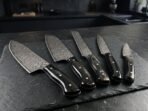 Damascus chef, santoku, bread, utility and paring knives with black horn handles fanned out on a slate board