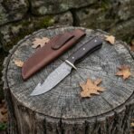 Damascus gut hook hunting knife with dark wenge wood handle on a tree stump beside its brown leather sheath and oak leaves