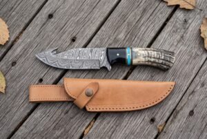 Damascus gut hook knife with ram horn and buffalo horn handle and turquoise spacer above its tan leather sheath on grey planks