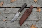 Damascus hunting knife with wenge wood handle seen from above on weathered boards, brown leather sheath and dry leaves