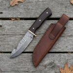 Damascus hunting knife with wenge wood handle seen from above on weathered boards, brown leather sheath and dry leaves