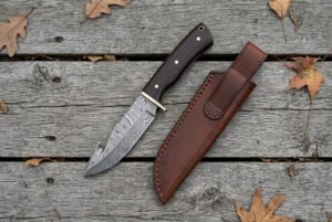 Damascus hunting knife with wenge wood handle seen from above on weathered boards, brown leather sheath and dry leaves