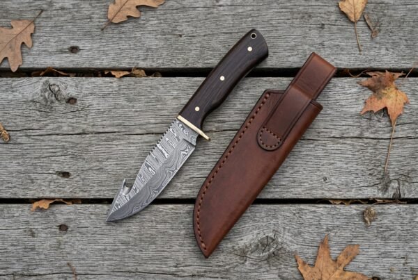 Damascus hunting knife with wenge wood handle seen from above on weathered boards, brown leather sheath and dry leaves