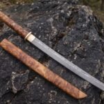 Damascus katana with burl wood handle and pale collar laid beside its burl wood sheath on a black boulder