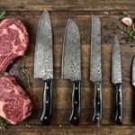 Damascus steel five piece kitchen knife set with black horn handles laid out beside raw steaks, garlic and herbs on a wooden board