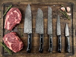 Damascus steel five piece kitchen knife set with black horn handles laid out beside raw steaks, garlic and herbs on a wooden board