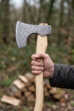 Damascus Viking bearded axe held upright in one hand by its birch wood handle in a woodland setting