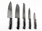 Five piece Damascus kitchen knife set with black horn handles and pins shown side by side on a plain white background