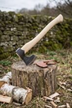 Hand forged bearded axe with birch wood handle, head sunk into a chopping stump beside its leather blade cover and a stone wall