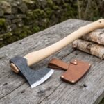 Hand forged bearded axe with birch wood handle on a weathered outdoor table next to its leather blade cover and split birch logs