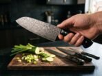 Hand holding a Damascus santoku knife with black horn handle over sliced cucumber and spring onion, more knives on the board