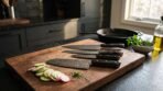 Damascus kitchen knife set with black horn handles resting on a walnut cutting board beside sliced cucumber and radish