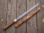 Straight Damascus katana and its olive burl wood sheath laid side by side on weathered planks with oak leaves