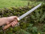 Straight Damascus katana with olive burl wood handle held out in one hand in front of a green field wall