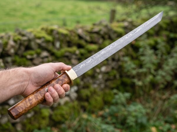 Straight Damascus katana with olive burl wood handle held out in one hand in front of a green field wall