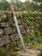 Straight Damascus katana with olive burl wood handle leaning against a mossy dry stone wall