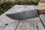 Damascus steel skinner knife blade in close side view, layered pattern and curved belly over weathered wood