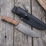 Damascus steel skinner knife with stabilized wood handle on grey boards from above, beside a black leather sheath