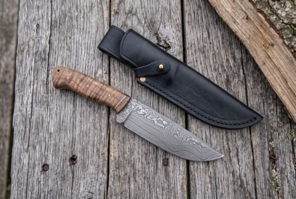 Damascus steel skinner knife with stabilized wood handle on grey boards from above, beside a black leather sheath