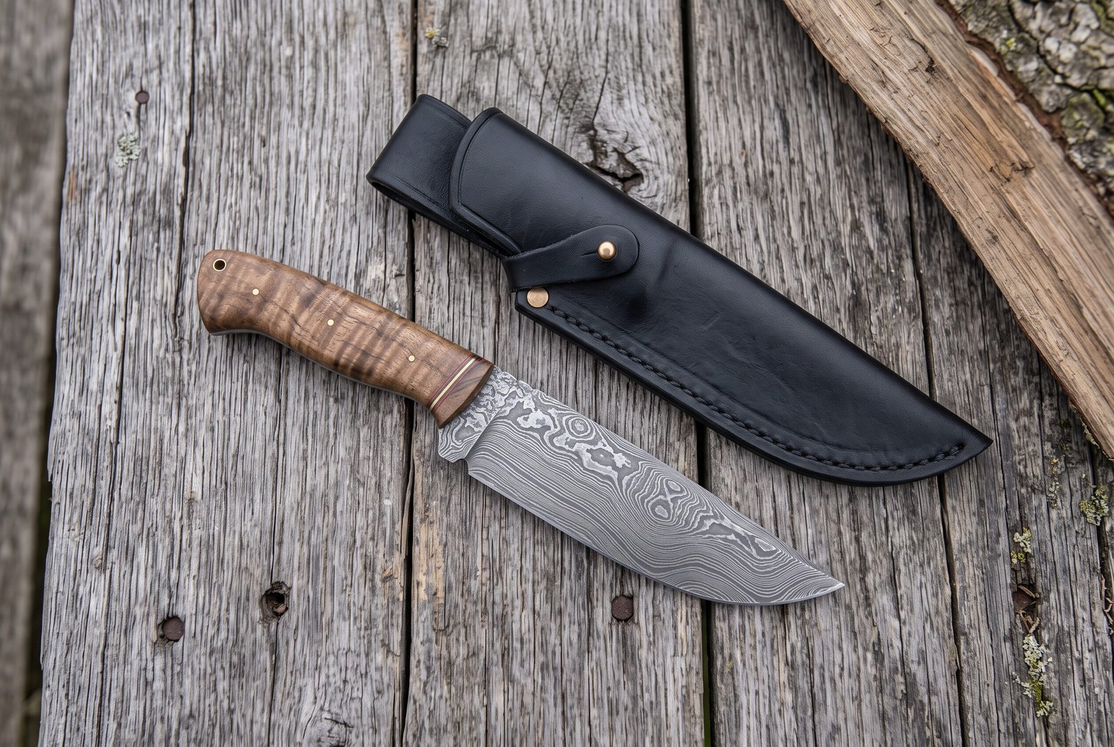 Damascus steel skinner knife with stabilized wood handle on grey boards from above, beside a black leather sheath Damascus steel skinner knife with stabilized wood handle on grey boards from above, beside a black leather sheath
