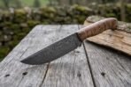 Damascus steel skinner knife with stabilized wood handle resting on a log, blade angled toward the camera