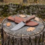 Damascus steel skinner knife with walnut handle on a tree stump across its tan leather sheath with oak leaves