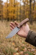 Full tang Damascus skinner knife with walnut wood handle held in one hand in an autumn forest