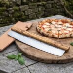 Stainless steel rocking pizza cutter with olive wood handle on a wooden board beside a sliced pizza and leather cover
