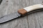 Damascus folding knife close up of the white bone handle and hardwood bolster with the patterned blade edge