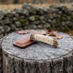 Damascus folding pocket knife with natural bone handle and stag bolsters closed on a tree stump with a brown leather sheath
