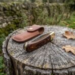 Damascus folding pocket knife with pakka wood handle shown closed on a tree stump beside its brown leather sheath