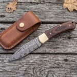 Damascus folding pocket knife with pakka wood handle open on old boards from above, leather sheath and oak leaves around it
