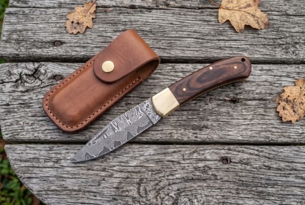 Damascus folding pocket knife with pakka wood handle open on old boards from above, leather sheath and oak leaves around it