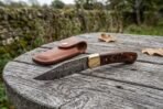 Damascus folding pocket knife with pakka wood handle lying open on a tree stump, brown leather sheath behind it