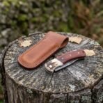 Damascus folding pocket knife with rosewood handle and stag bolster lying closed on a stump next to its leather sheath