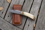 Damascus pocket knife with bone handle open beside its brown leather belt sheath on weathered planks, seen from above