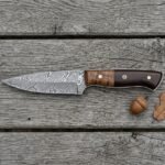 Damascus skinner knife with walnut and dark pakka wood handle lying on grey boards beside an acorn and oak leaf