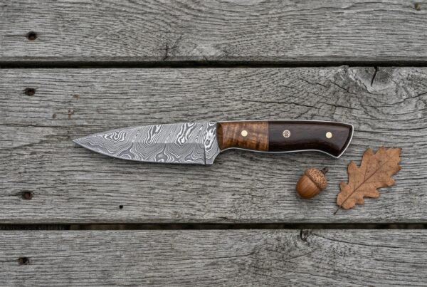 Damascus skinner knife with walnut and dark pakka wood handle lying on grey boards beside an acorn and oak leaf