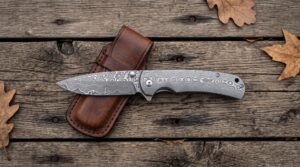 Open Damascus steel folding pocket knife laid on a brown leather sheath on grey wooden boards with autumn oak leaves