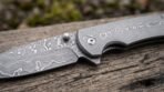 Damascus steel folding pocket knife close up of the patterned blade, pivot and flipper resting on weathered wood