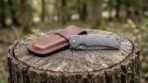 Damascus steel folding pocket knife closed beside its brown leather slip sheath on top of a cut tree stump in a forest