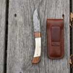 Overhead view of the open Damascus knife with bone and wood handle beside its brown leather belt pouch