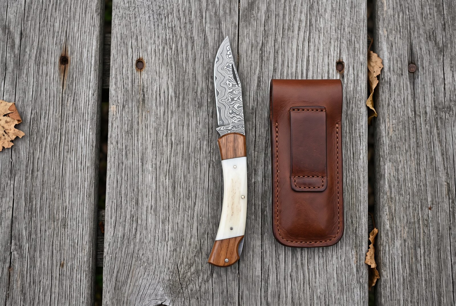 Overhead view of the open Damascus knife with bone and wood handle beside its brown leather belt pouch Overhead view of the open Damascus knife with bone and wood handle beside its brown leather belt pouch