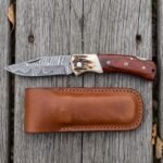 Top view of the open Damascus folding knife with stag bolster and rosewood handle above its brown leather sheath on wood