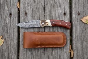 Top view of the open Damascus folding knife with stag bolster and rosewood handle above its brown leather sheath on wood