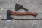 Bearded axe with rosewood handle lying on grey wooden boards beside its dark brown leather blade cover and wood shavings