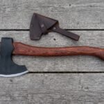 Bearded axe with rosewood handle lying on grey wooden boards beside its dark brown leather blade cover and wood shavings