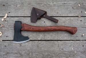 Bearded axe with rosewood handle lying on grey wooden boards beside its dark brown leather blade cover and wood shavings